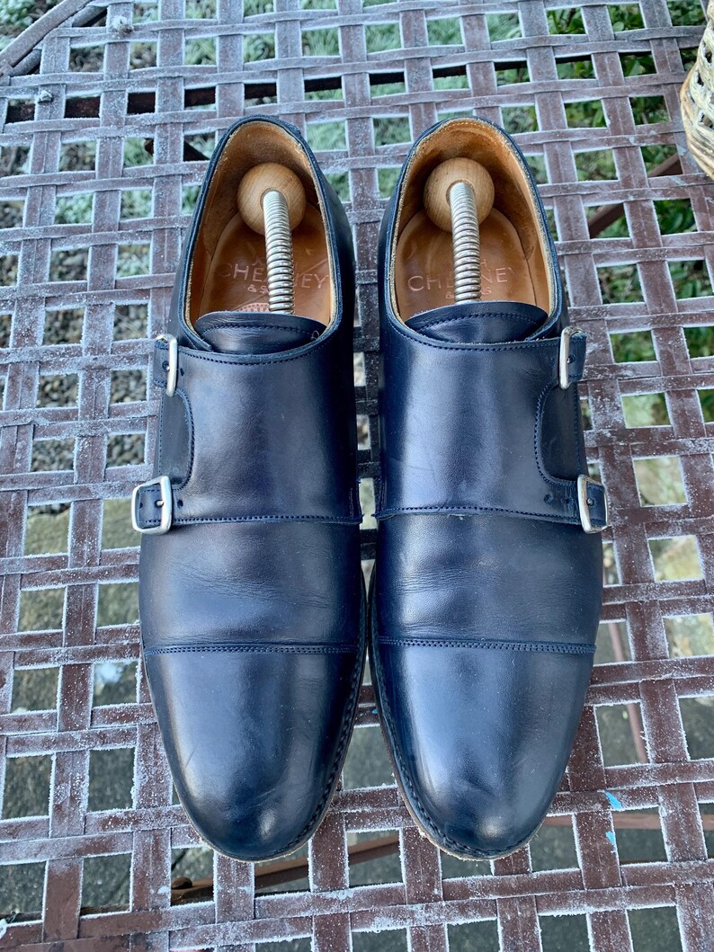 May include: A pair of navy blue leather double monk strap shoes with silver buckles. The shoes have wooden shoe trees inside. The word "CHEY" is visible on the inner lining of one shoe.