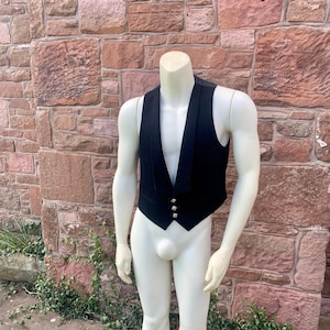 May include: A black waistcoat with a V-neck and three silver buttons. The garment is displayed on a headless mannequin against a red brick wall. The waistcoat is a formal garment.