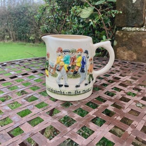 Könnte beinhalten: Cremefarbener Keramikkrug mit Henkel und Ausguss. Der Krug hat ein Reliefdesign von Figuren in farbenfroher Kleidung und den Text "Widecombe-in-the-Moor". Der Krug steht auf einem braunen Metalltisch.