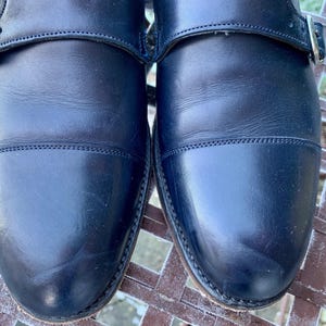 May include: Close-up of a pair of black leather dress shoes. The shoes have a cap-toe design and a buckle closure. The leather appears to be polished and in good condition. The shoes are sitting on a metal surface.
