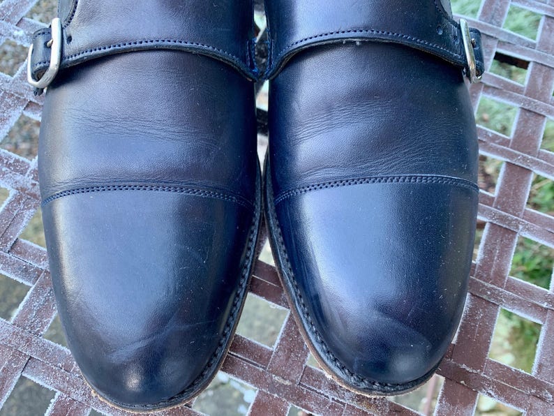 May include: Close-up of a pair of black leather dress shoes. The shoes have a cap-toe design and a buckle closure. The leather appears to be polished and in good condition. The shoes are sitting on a metal surface.
