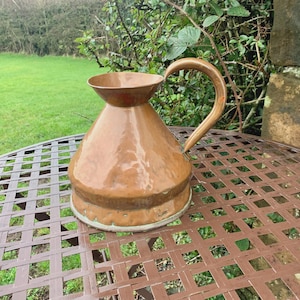 Puede incluir: Una jarra de cobre con un asa curva y un pico acampanado. La jarra tiene un cuerpo bulboso y se asienta sobre una mesa redonda de metal estampada. La jarra está al aire libre, con hierba verde y follaje en el fondo.