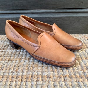 May include: A pair of tan leather slip-on shoes with a low heel. The shoes have a rounded toe and are stitched with dark thread around the edges and seams. The heels are dark brown and the shoes are on a woven rug.