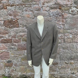 May include: A brown blazer with a subtle vertical stripe pattern. The blazer has a notched lapel, two front pockets, and a two-button closure. The jacket is displayed on a mannequin with white gloves and pants.