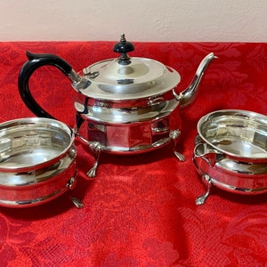 Puede incluir: Un juego de té de plata con tetera, jarra de crema y azucarero. La tetera tiene un asa negra y un pomo de plata en la tapa. La jarra de crema y el azucarero tienen tres patas cada uno.