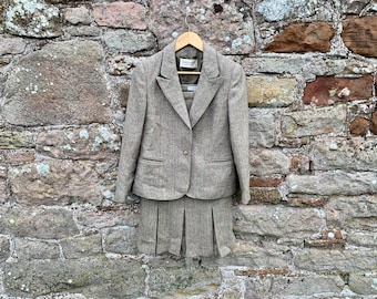 Op de afbeelding: Een tweed tweedelig pak, bestaande uit een blazer en een geplooide rok, hangt aan een houten hanger. Het pak heeft een lichtbruine kleur met een subtiel visgraatpatroon. De blazer heeft een sluiting met één knoop en twee voorzakken.