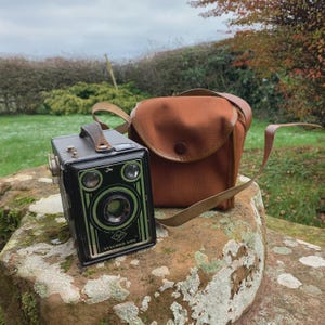 May include: A vintage black camera with green detailing and the text "SYNCHRO BOX" alongside a brown camera bag with a leather strap. The camera rests on a large rock in an outdoor setting.