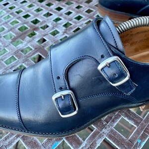 May include: Close-up of a pair of navy blue leather double monk strap shoes. The shoes feature silver buckles and stitching details. A shoe tree is inserted in the shoe. The shoes are on a metal surface.