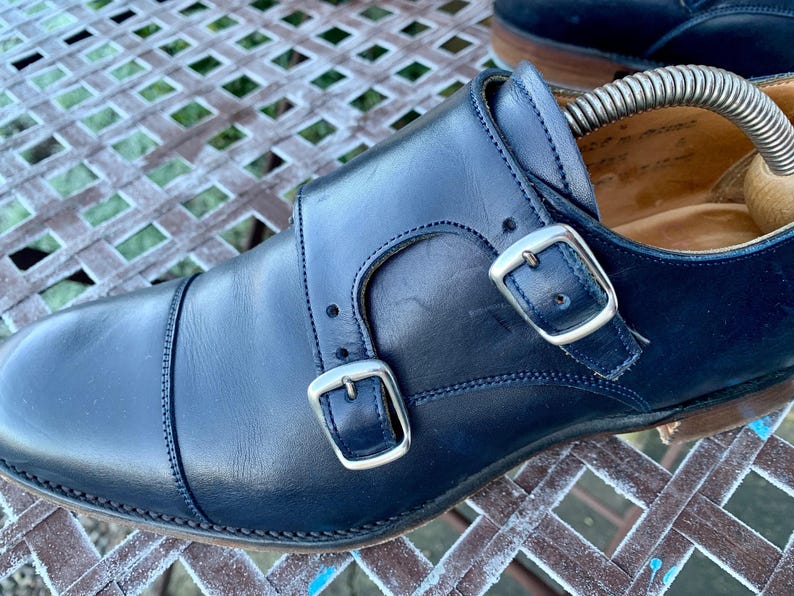 May include: Close-up of a pair of navy blue leather double monk strap shoes. The shoes feature silver buckles and stitching details. A shoe tree is inserted in the shoe. The shoes are on a metal surface.