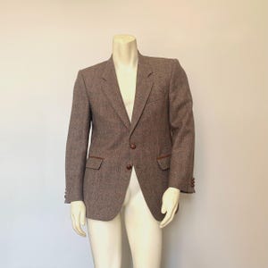 May include: A brown tweed blazer with a two-button closure, notched lapels, and two front pockets. The blazer has brown leatherette trim on the pockets and lapels. The sleeves have three buttons at the cuffs. The blazer is displayed on a mannequin.