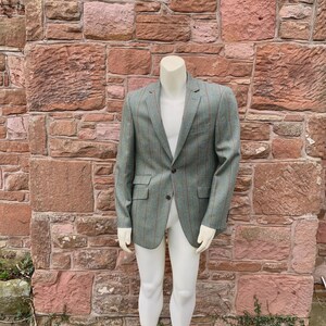 May include: A green blazer with red pinstripes is displayed on a headless mannequin. The blazer has two front pockets and a single-button closure. The mannequin is wearing white pants. The background is a brick wall.