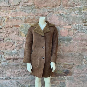 Peut inclure: Un manteau en peau de mouton marron avec une fermeture à boutons et un col en peau de mouton. Le manteau est présenté sur un mannequin. Le manteau a un design classique et semble être fait d'une matière douce et chaude. Le manteau est de longueur moyenne.