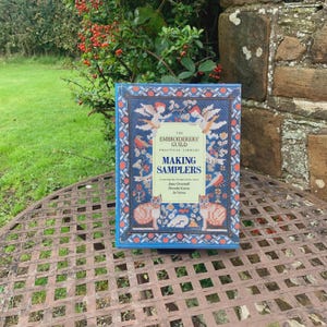 Może przedstawiać: Książka zatytułowana "MAKING SAMPLERS" z The Embroiderers' Guild Practical Library. Okładka ma niebieską obwódkę z haftowanymi ptakami, kotami i motywami kwiatowymi. Książka leży na metalowym stole na zewnątrz, z zielenią i kamiennym murem w tle.