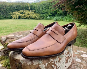 Puede incluir: Un par de mocasines penny de cuero marrón claro con un diseño clásico. Los zapatos se exhiben sobre una superficie de piedra, mostrando su acabado pulido y su elegante silueta. Los mocasines presentan un estilo sin cordones con una correa en el empeine.