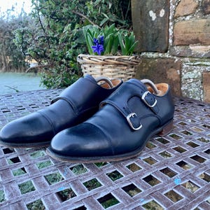 May include: A pair of black leather double monk strap shoes with silver buckles. The shoes are displayed on a metal table with a basket of purple flowers in the background. The shoes have a classic, formal design.