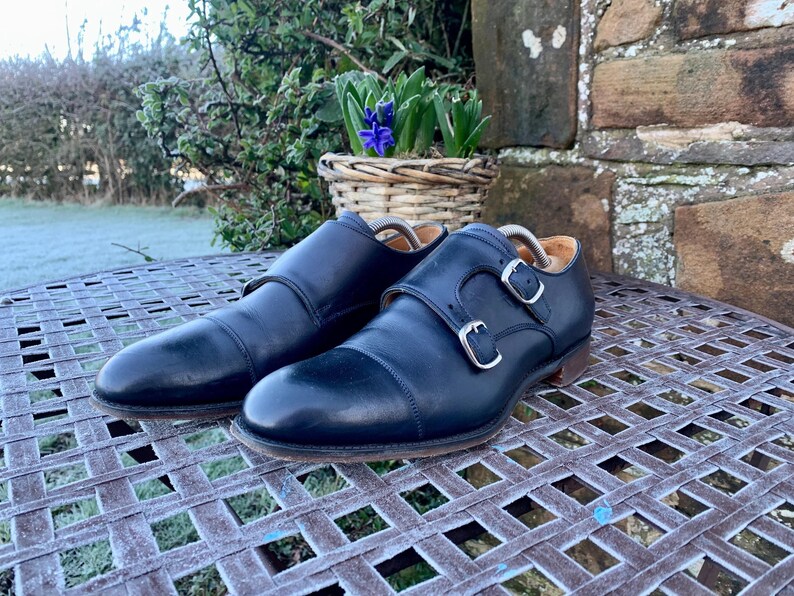 May include: A pair of black leather double monk strap shoes with silver buckles. The shoes are displayed on a metal table with a basket of purple flowers in the background. The shoes have a classic, formal design.