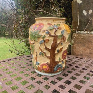 May include: A beige ceramic vase with a detailed relief design of a tree and flowers. The vase has a wide mouth and a light blue base. The tree is brown, and the flowers are red, yellow, and blue. The vase is on a brown metal table.