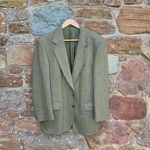 Op de afbeelding: Een groene tweed blazer met een sluiting met twee knopen en een revers hangt aan een houten hanger. De jas heeft twee voorzakken en een subtiel patroon. De blazer wordt gepresenteerd tegen een stenen muur.