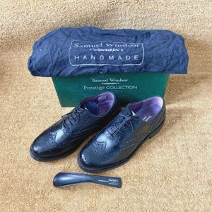 Può includere: Scarpe brogue in pelle nera con dettagli intricati, lacci e un calzascarpe. Le scarpe sono esposte con un sacchetto antipolvere blu scuro Samuel Windsor Handmade e una scatola verde con l'etichetta "Samuel Windsor Prestige Collection".