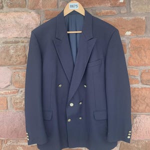 May include: A navy blue double-breasted blazer with gold-colored buttons. The blazer is hanging on a wooden hanger against a brick wall. The blazer has two rows of buttons and pockets on each side.