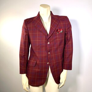 Pode incluir: Um blazer bordô com um padrão xadrez em tons de azul e dourado. O blazer tem fecho de três botões, um bolso no peito com um lenço de bolso estampado e dois bolsos frontais. O blazer é exibido em um manequim.