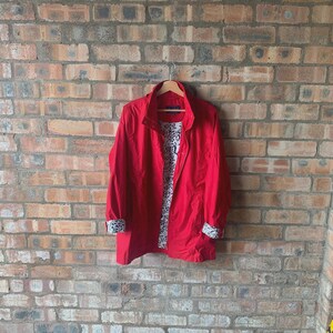 Puede incluir: Una chaqueta roja brillante con cuello alto y cierre de cremallera, colgada de una percha de madera. El interior y los puños de la chaqueta presentan un estampado floral blanco y negro. La chaqueta se exhibe contra una pared de ladrillos.