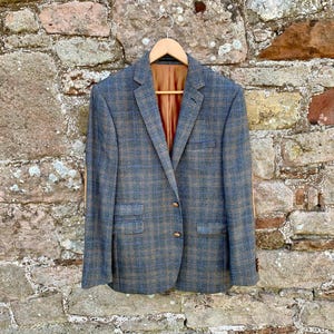 May include: A blue and brown plaid blazer with a brown interior lining hangs on a wooden hanger. The blazer has two front pockets and a single button closure. The background is a weathered stone wall.
