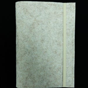 Könnte beinhalten: Eine hellbeige Filz-Buchhülle mit passendem Gummibandverschluss. Die Hülle hat eine gesprenkelte Textur und ist rechteckig. Das Gummiband verläuft vertikal entlang der rechten Seite der Hülle.