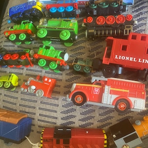 May include: A collection of colorful toy trains, including a red fire truck, a red caboose with "Lionel Lines" text, and several green and blue locomotives. The trains are arranged on a patterned surface.
