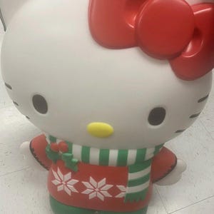 May include: Large white Hello Kitty figure with a red bow, black eyes, and a yellow nose. It wears a red sweater with white snowflake designs and a green and white striped scarf. The figure is a festive holiday decoration.