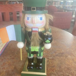 May include: A wooden leprechaun nutcracker wearing a green top hat and coat, holding an Irish flag and a pot of gold. The nutcracker has a white face, a long, tan beard, and black boots. The base is green with gold trim.