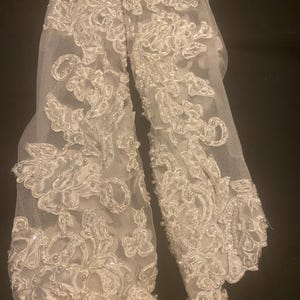 May include: A pair of white, long, fingerless gloves. The gloves are made of a sheer, lacy fabric with intricate floral embroidery and small, pearl-like beads. The design is symmetrical, with detailed patterns.