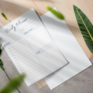 May include: A white daily planner with sections for schedule, today's goals, and notes. The planner is on a wooden surface with green leaves.