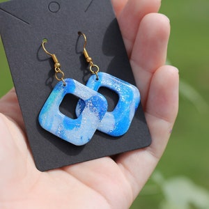 May include: A pair of blue and white square earrings with gold hooks. The earrings have a marbled effect and glitter embedded in the resin.