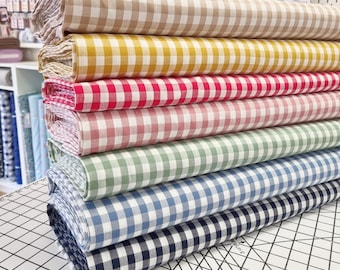 Puede incluir: Un rollo de tela en varios colores y patrones de cuadros. Los rollos están apilados en una estera de corte blanca con líneas de cuadrícula negras. Los colores de la tela son beige, amarillo, rojo, rosa, verde, azul y negro.