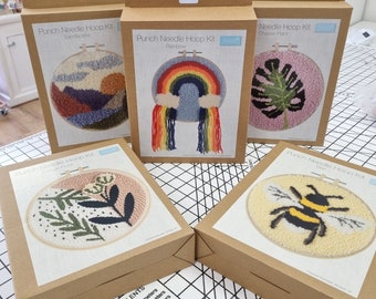May include: Five cardboard boxes with punch needle kits. The kits feature different designs including a landscape, rainbow, cheese plant, bee, and floral design. Each box has a clear plastic window showing the design of the kit.