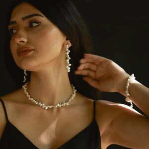 A woman in a black dress elegantly showcases a sterling silver keshi pearl jewelry set, featuring a delicate pearl necklace, long dangle earrings, and a matching bracelet. The organic shape of the pearls adds a natural and refined charm to her sophisticated look.