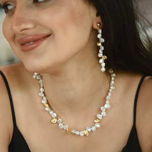 A woman with long dark hair wearing a handmade keshi pearl jewelry set, featuring a dainty necklace with white freshwater pearls and gold-plated details, paired with matching cascading pearl earrings. The organic design exudes elegance and sophistication.