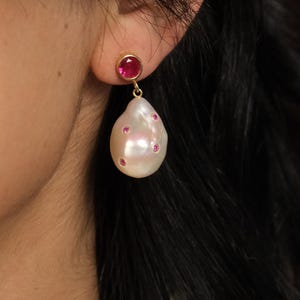 Puede incluir: Primer plano de un pendiente de oro con una pequeña gema roja redonda y una gran perla blanca en forma de lágrima con gemas rojas más pequeñas. El pendiente se lleva en la oreja de una persona.