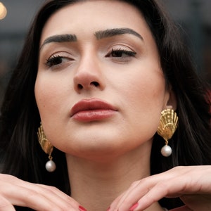 Op de afbeelding: Een vrouw met lang donker haar draagt een donkerblauwe top met een parelketting. Ze draagt gouden oorbellen met een parel. Haar nagels zijn rood gelakt.