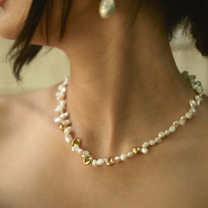 A close-up of a woman wearing a handmade keshi pearl jewelry set, featuring a delicate necklace with irregular white freshwater pearls and gold-plated accents, paired with an elegant baroque pearl drop earring. The organic shapes add a refined, timeless charm.