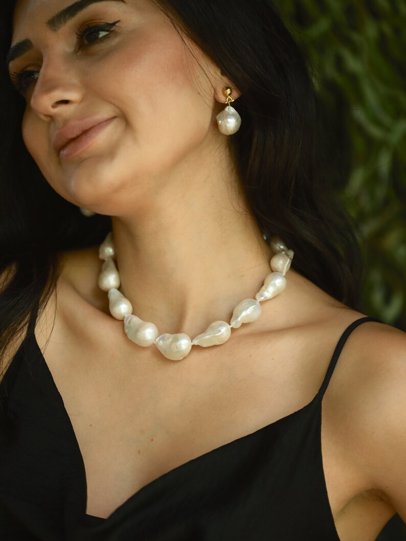 A woman wearing a chunky baroque pearl necklace and a matching drop earring smiles softly. The bold, organic shapes of the pearls stand out against her black dress. Her dark hair and the leafy green background add a natural, effortless vibe to the look.