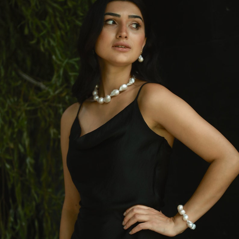 A woman in a sleek black dress shows off her bold baroque pearl set, including a chunky necklace, matching bracelet, and drop earrings. She stands confidently with one hand on her hip, looking off to the side.