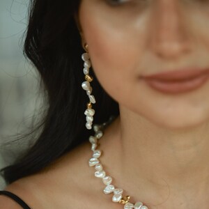 A close-up of a woman wearing a delicate keshi pearl jewelry set, featuring a handcrafted necklace adorned with organic-shaped freshwater pearls and gold-plated accents, complemented by a long, cascading pearl earring for a timeless, elegant look.
