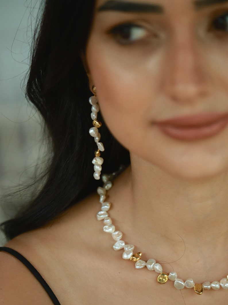 A close-up of a woman wearing a delicate keshi pearl jewelry set, featuring a handcrafted necklace adorned with organic-shaped freshwater pearls and gold-plated accents, complemented by a long, cascading pearl earring for a timeless, elegant look.