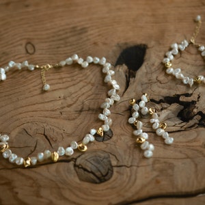 A handcrafted keshi pearl jewelry set displayed on a rustic wooden surface, featuring a necklace, a matching bracelet, and elegant dangle earrings. The set is adorned with natural white freshwater pearls and subtle gold-plated accents, offering a delicate and timeless aesthetic.