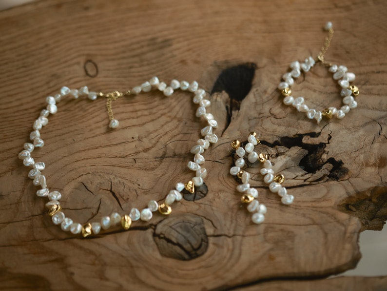 A handcrafted keshi pearl jewelry set displayed on a rustic wooden surface, featuring a necklace, a matching bracelet, and elegant dangle earrings. The set is adorned with natural white freshwater pearls and subtle gold-plated accents, offering a delicate and timeless aesthetic.