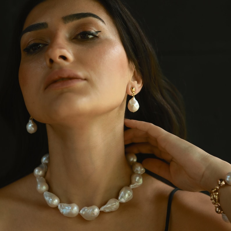 A woman is shown wearing an elegant set of jewelry featuring large, white baroque pearls. The necklace and drop earrings highlight the organic shapes and natural luster of the pearls, adding a bold yet refined touch to her look. Her hand gently touches her neck, creating a graceful and poised moment.