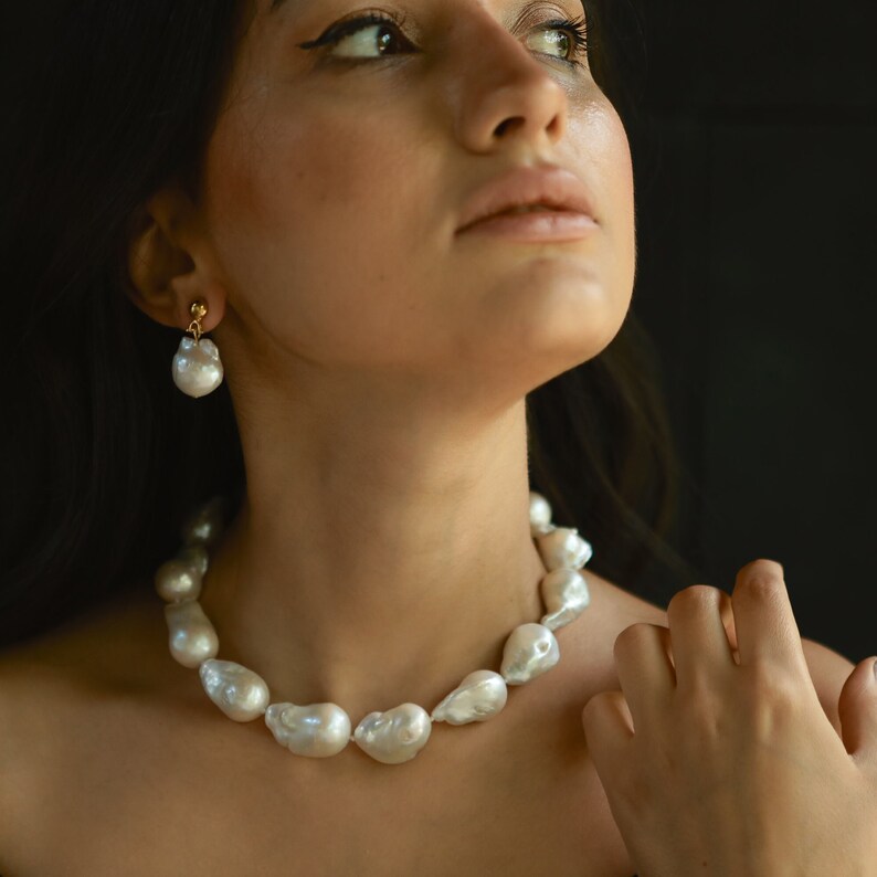 A woman is wearing a bold baroque pearl necklace with chunky, white freshwater pearls. Her matching drop earrings add a touch of elegance to the look. She gazes slightly upward with a soft expression, standing against a dark background that makes the pearls stand out beautifully.