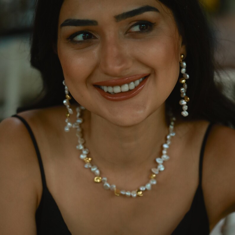 A woman with dark hair and a warm smile wears a handmade pearl jewelry set featuring a white keshi pearl necklace and dangling pearl earrings. The organic pearl shapes add a unique, elegant touch, making this set perfect for weddings, special occasions, or everyday luxury.